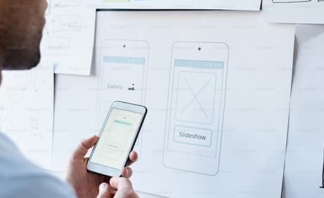 Close-up of a person using a mobile phone with UX wireframes on a whiteboard in the background.